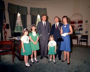Three young girls in green dresses, a suited man, and a woman in blue pose with president John F. Kennedy in the Oval Office with large windows, desk, and bookshelves.