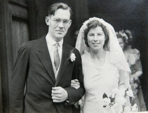 A black and white photo of a smiling bride in a veil with bouquet and groom in glasses, suit, and boutonnière on their wedding day during World War II.