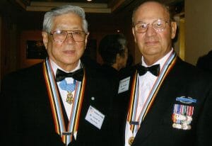 Two elderly men in tuxedos with glasses, bow ties, medals, and ribbon lanyards stand side by side, smiling at the camera.