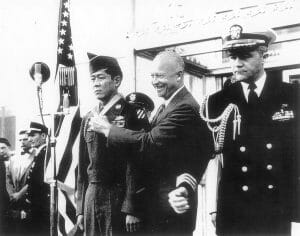 A military officer is awarded a medal by a smiling President Eisenhower as another uniformed officer stands nearby; U.S. flag and microphone behind.