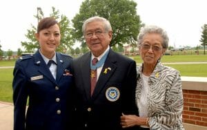 Three people stand outdoors smiling: a young woman in an Air Force uniform, an older man with a medal, and an older woman.