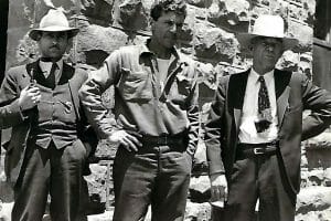 Three men pose seriously before a stone wall; two wear suits and hats, the center man wears a collared shirt and pants, in sunlight.