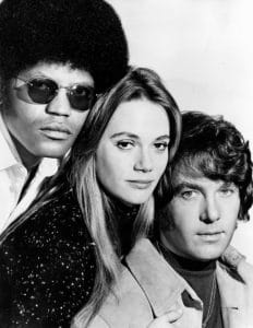 Three young adults pose in a black-and-white photo: a man with sunglasses and an afro, a woman with long hair, and a man with wavy hair.