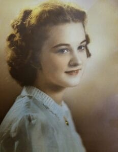 A young woman with wavy brown hair, in a white blouse with a ruffled collar and brooch, smiles gently, looking to the side. It is a hand-colored formal portrait from the early 1940s.