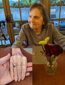 An elderly woman with brown hair smiles at a table near red and yellow flowers. Inset: her wrinkled hand rests in a man's hand.