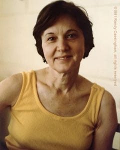 A woman with short dark hair in a yellow sleeveless top smiles at the camera. Neutral background, warm light.