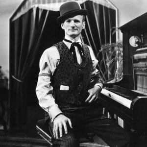 A man in a bowler hat, vest, and striped pants sits at an upright piano. A curtain and plant are behind him. Black and white photo.