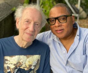 Two men sit closely outdoors, both smiling at the camera—one older with white hair and a jazz shirt, one younger in glasses.