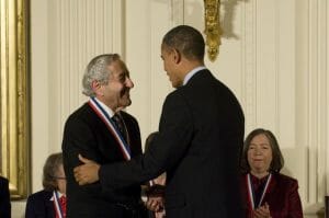 A man wearing a medal shakes hands and smiles with President Obama at an indoor ceremony as others clap in the background.