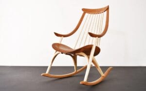 A modern wooden rocking chair with a curved seat, tall spindle backrest, and light and dark wood tones on a white background.