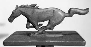 A wooden sculpture of a galloping horse in mid-stride sits on a rectangular base against a plain background, showing detailed features.