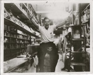Two young men in a store; one sits on the other's shoulders, smiling with arms raised, while shelves of products and a ladder are visible in the background. The image is in black and white.