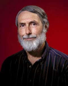 An older man with gray hair and a full white beard, wearing a dark striped shirt, poses against a deep red background. He is facing forward and smiling slightly.