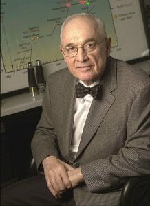 An older man in glasses and a bow tie sits in an office chair, wearing a brown checked suit. A scientific chart is lit behind him.