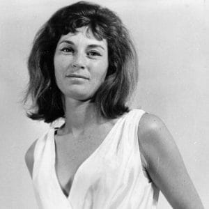 A woman with shoulder-length dark hair in a sleeveless white top stands before a plain light background, smiling slightly at the camera.