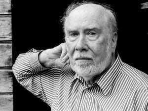 An older man with a white beard and mustache in a striped shirt stands before a dark background, hand near neck, facing the camera.