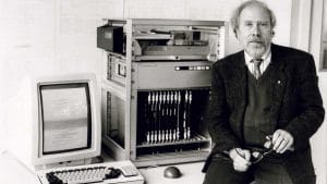 A man in a suit sits next to an early computer system with a monitor showing text, a keyboard, mouse, and rack-mounted components.