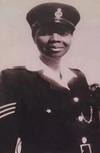 Black-and-white photo of a smiling woman in police uniform and cap, with sleeve insignia and a white shirt under her dark jacket.
