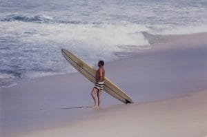 A man in striped swim trunks stands on a sandy beach with a long surfboard, gazing at ocean waves rolling toward the shore.