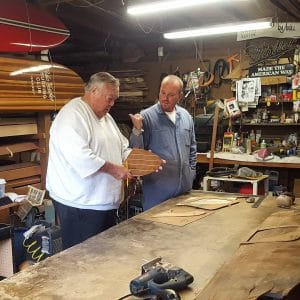 Two men in a busy workshop, one holding wood, the other pointing. Surfboards hang on the wall amid tools and scattered materials.