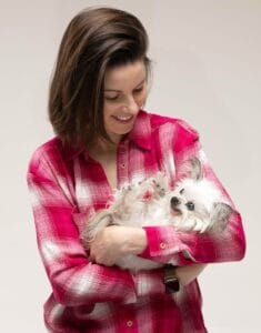 A smiling woman in a pink plaid shirt holds a fluffy white dog with gray ears, both looking happy against a plain background.