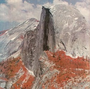 A false-color image showing Half Dome’s granite dome towering over a forest with reddish leaves beneath a partly cloudy sky in Yosemite National Park.