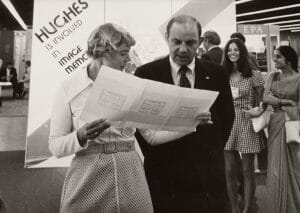 A woman holds a large sheet of paper and speaks with a man at an event. Behind them is a display about image memory, with other people in the background.