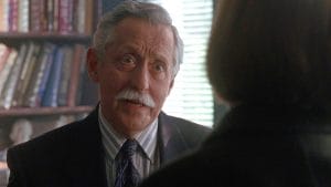 An older man with gray hair and a mustache in a suit and tie talks to someone seen from behind, in an office with bookshelves.