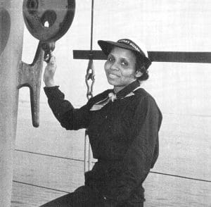 A woman in a U.S. Coast Guard uniform and hat smiles while sitting on a ship, holding onto a large wheel or pulley, with the ocean visible in the background.