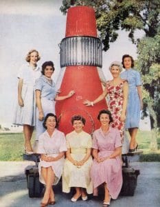 Seven women in 1960s dresses smile and pose outdoors before a red model spacecraft capsule, with green grass and trees behind them.