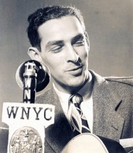 A man in a suit and tie sits behind a vintage WNYC microphone, smiling slightly while holding a guitar. The photo is black and white, giving it a classic, mid-20th-century feel.