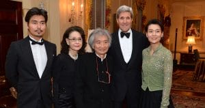 Five people in formal attire pose at an elegant indoor event with ornate decor, paintings, and warm lighting in the background.