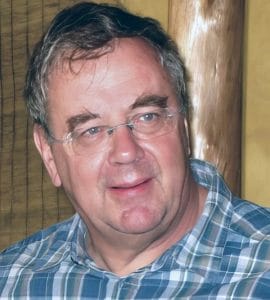 A middle-aged man with glasses and graying hair in a blue and white plaid shirt smiles, looking sideways in a warmly lit wood-toned room.
