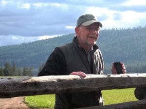 Paul Brainerd in a cap, glasses, and vest stands behind a wooden fence, smiling and holding a smartphone, with green hills and a cloudy sky in the background.