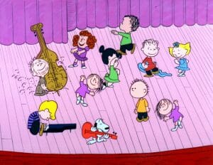 A frame from a 'Peanuts' TV special shows a group of animated children and a dog dance on a stage with a pink curtain, some playing instruments, in cartoon style.