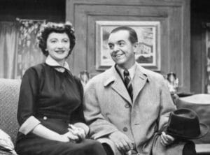 A woman in a dark dress and a man in a light coat holding a hat sit side by side on a couch, smiling. They appear to be on a vintage set with curtains and a framed painting in the background.