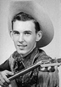 A smiling young man in a patterned western shirt and wide-brimmed cowboy hat holds a guitar. The photo is black and white.