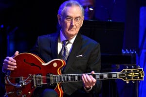 An older man in a suit sits on stage with a red electric guitar under blue lighting, prepared to play. No lectern is pictured.