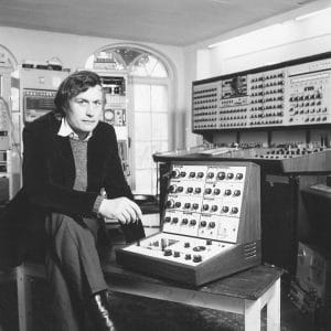 A man in a suit sits next to a modular synthesizer with lots of knobs in a vintage electronic music studio with large equipment.