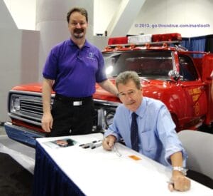 Two men at a convention: one is standing and smiling in a purple shirt, while the other sits at a table signing autographs. In the background, there is a vintage red fire truck.