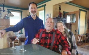 Three smiling adults pose together in a cozy, well-lit kitchen. Two cocktails rest on the counter. A man in a blue shirt stands, while an older man in plaid and a woman sit close by, all appearing cheerful.