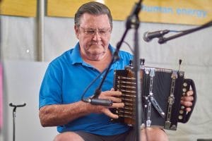 An older man in a blue shirt plays a button accordion, focused. A microphone is in front, with a blurred sign in the background.
