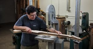 A man with glasses operates a bandsaw to cut a long wooden board in a workshop stocked with woodworking tools and machines.