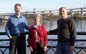 Three adults by a riverside railing, bridge and highways behind them; two men flank a woman. All wear casual clothes and smile.