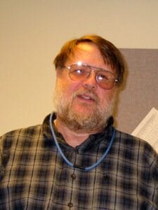 A man with light brown hair, a beard, and glasses is wearing a plaid shirt. He is standing indoors in front of a beige wall with a paper pinned up.