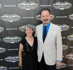 A woman in a black dress and a man in a light suit and blue shirt smile together before The Academy of Magical Arts backdrop.