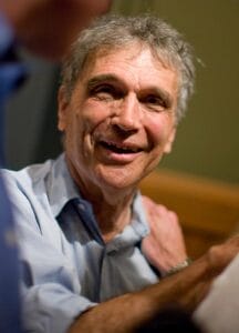 An older man with gray hair smiles warmly at the camera, wearing a light blue shirt indoors against a soft, blurred background.