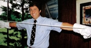 A smiling man in a blue shirt, tie, and white gloves stretches a long piece of material between his hands in an office with trees outside the window.