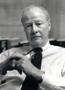 Black and white photo of an older man with light hair and mustache, in a white shirt and tie, holding glasses near bookshelves. His face bears a thoughtful expression.