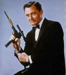 An actor in a black tuxedo and bow tie sits against a plain background, holding a large silenced gun, smiling slightly at the camera.